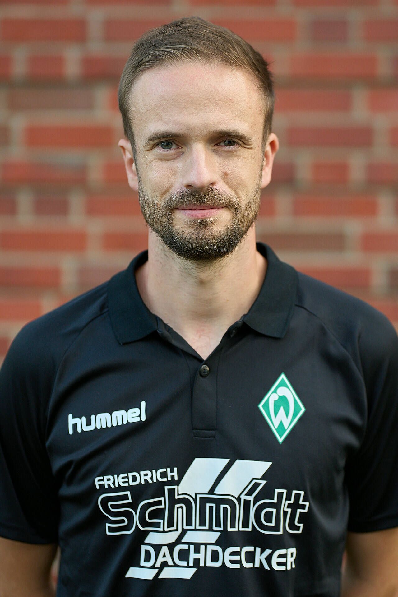 Lächelnder Mann im schwarzem Sportshirt mit Logos, der vor einer Backsteinmauer steht.
