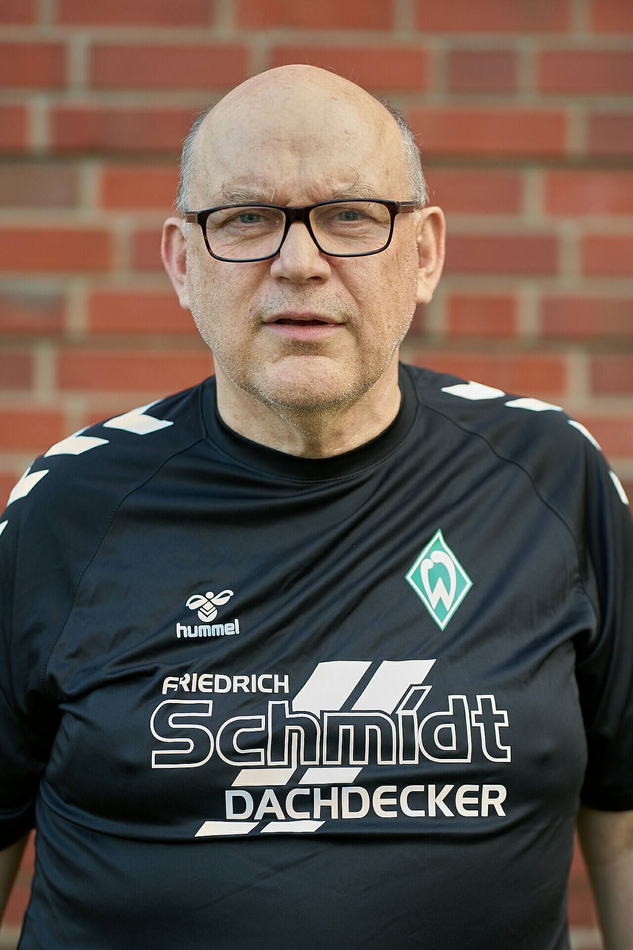 Lächelnder Mann mit Brille und schwarzem Sportshirt mit Logos, der vor einer Backsteinmauer steht.