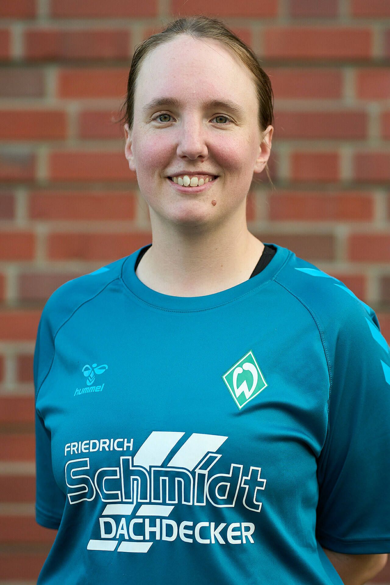 Lächelnde Frau im blauem Sportshirt mit Logos, die vor einer Backsteinmauer steht.