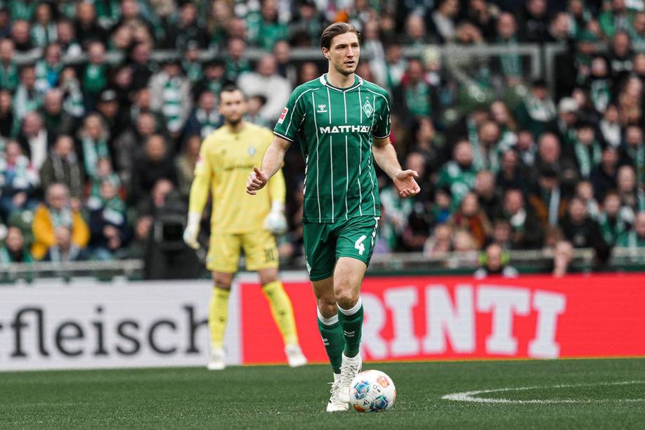 Fußballspieler in grün-weiß gestreiftem Trikot dribbelt den Ball während eines Spiels auf einem Feld, im Hintergrund sind ein Torwart und Zuschauer zu sehen.