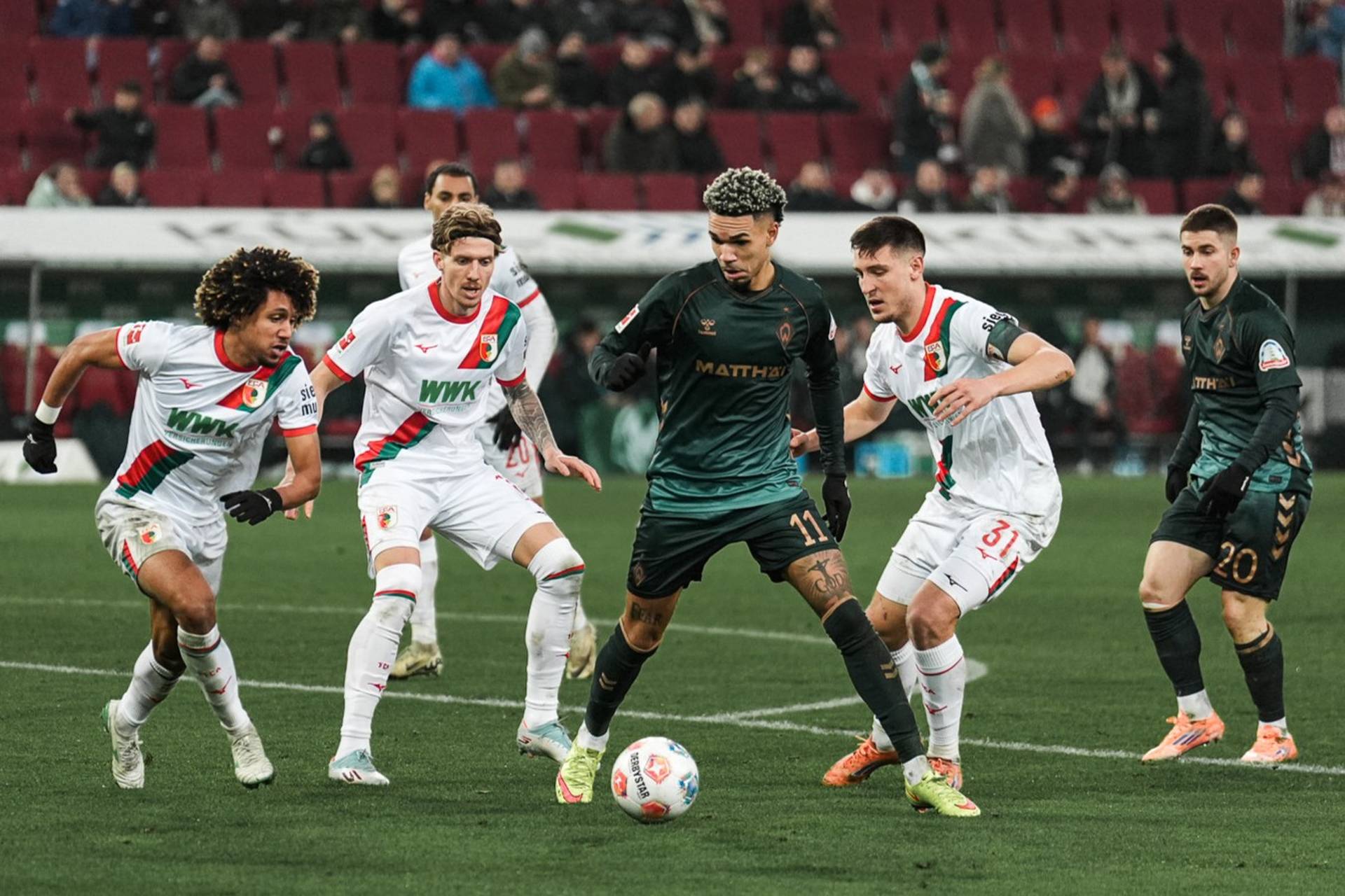 Justin Njinmah mit Ball am Fuß umzingelt von mehreren Augsburgern.