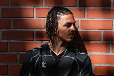 Kenny Quetant in black clothing leans against a sunlit brick wall with his arms crossed.