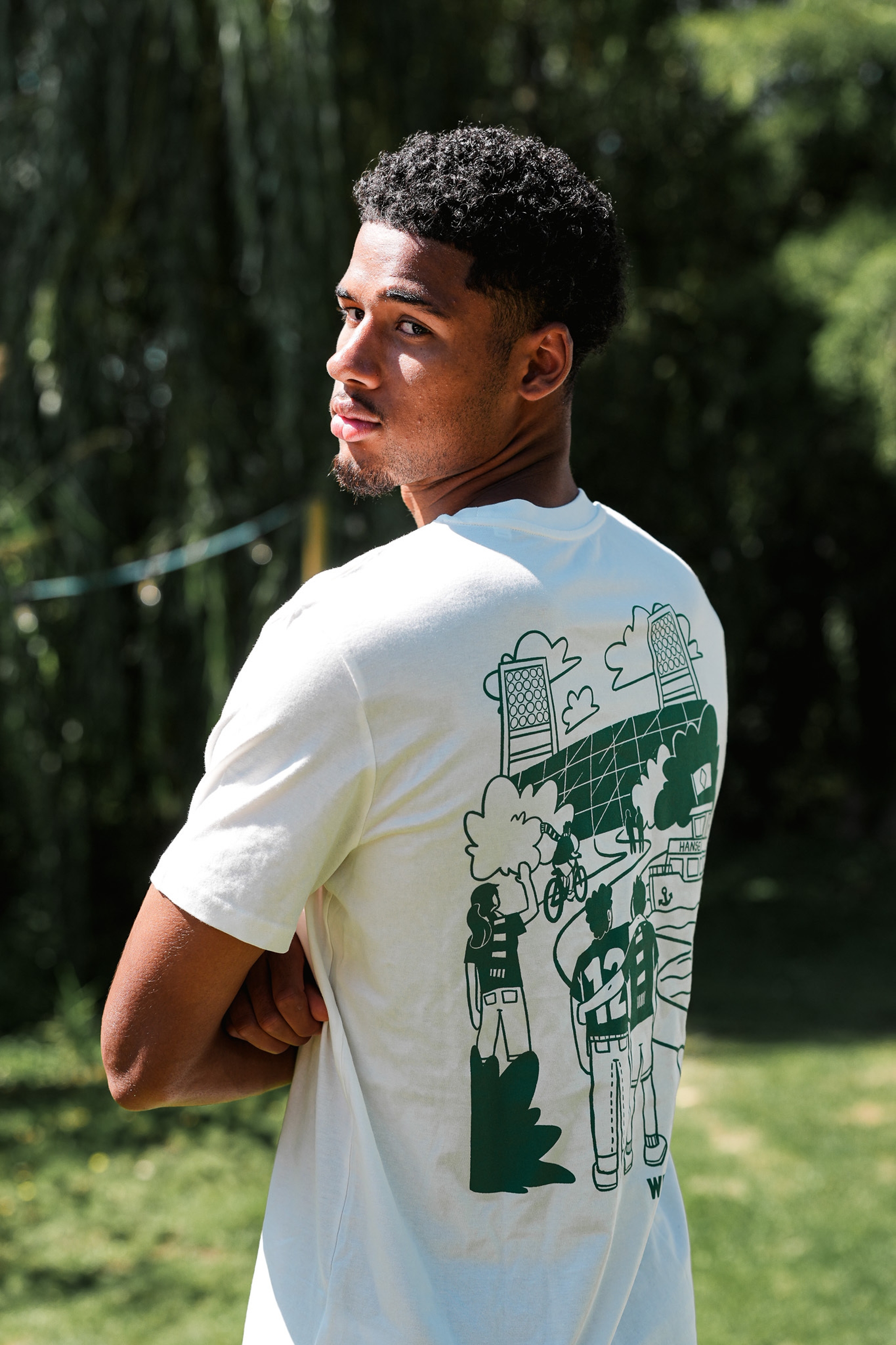 Eine Person mit lockigem Haar blickt zurück; sie trägt ein weißes T-Shirt mit einem grünen Motiv auf dem Rücken und steht draußen im Sonnenlicht.
