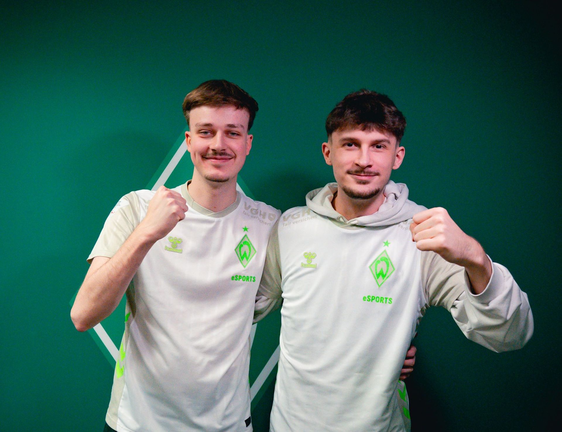Zwei Männer in eSPORTS Trikots posieren gemeinsam, lächeln und recken vor einem grünen Hintergrund triumphierend die Fäuste in die Höhe.