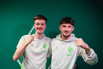 Zwei Männer in eSPORTS Trikots posieren gemeinsam, lächeln und recken vor einem grünen Hintergrund triumphierend die Fäuste in die Höhe.
