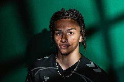 Ein junger Sportler mit Zöpfen steht mit einem Fußball in der Hand vor einem grünen Hintergrund und trägt ein schwarzes Sporttrikot.