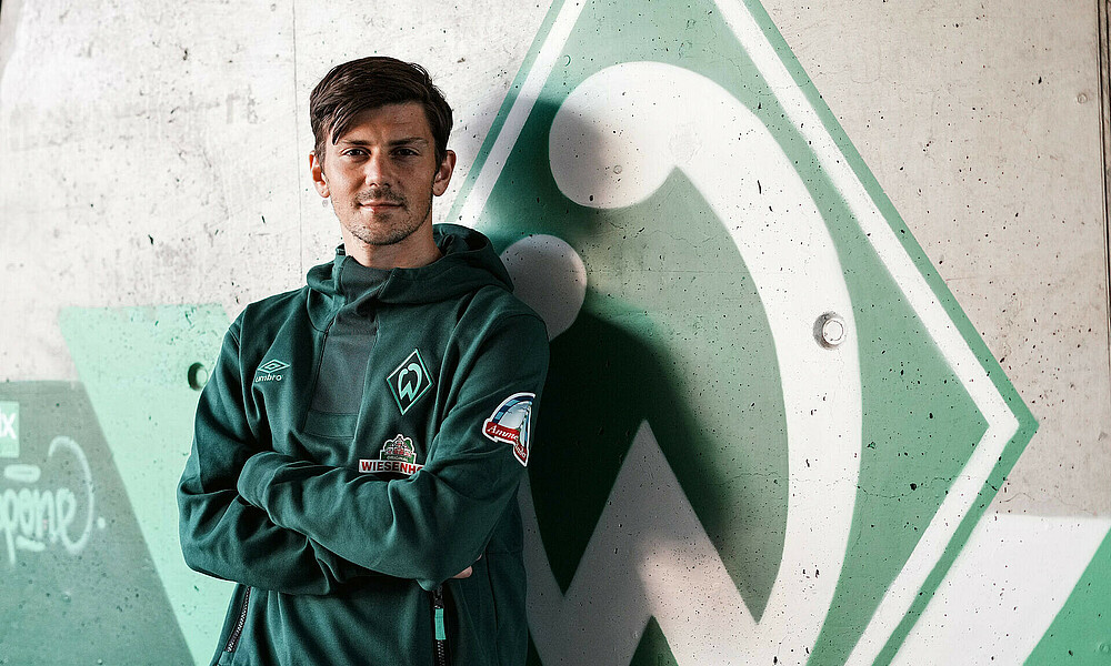 Dawid Kownacki lehnt im Werder-Pullover vor einer grauen Wand mit Werder-Raute.