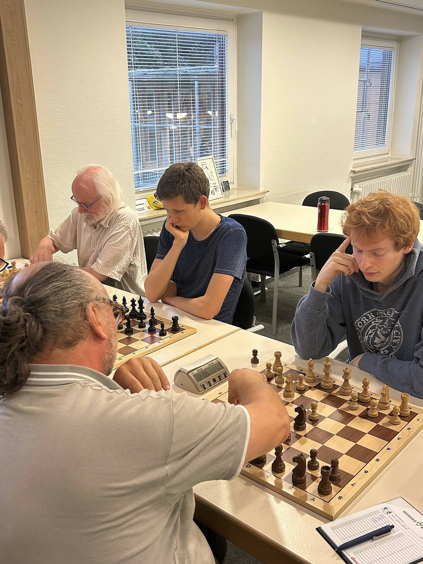 Vier Personen spielen intensiv Schach an einem Tisch in einem gut beleuchteten Raum, mit Schachbrettern und Uhren, und einem Fenster im Hintergrund.