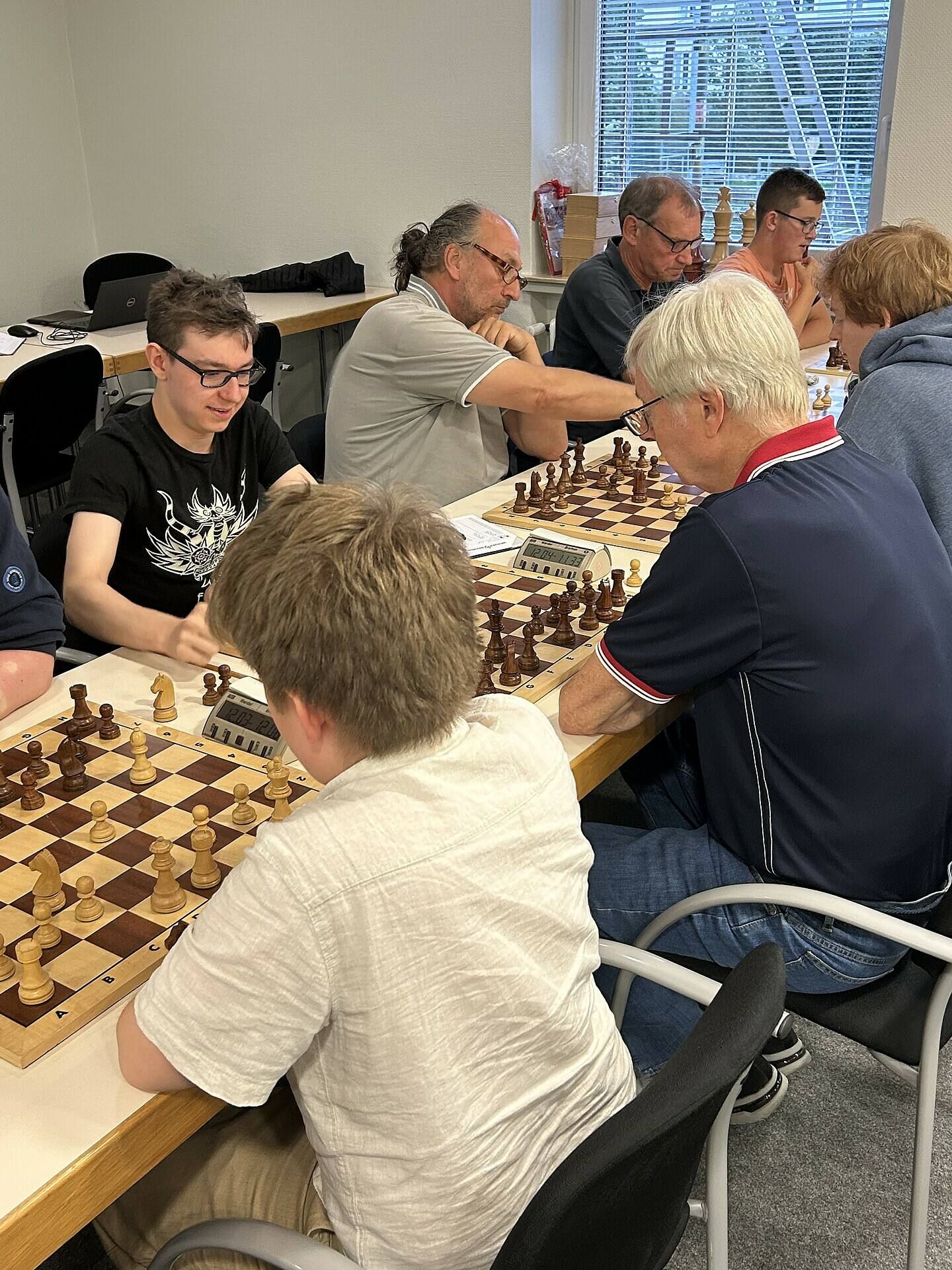 Menschen unterschiedlichen Alters spielen Schach an einem langen Tisch in einem Raum, in den durch ein Fenster Tageslicht fällt.