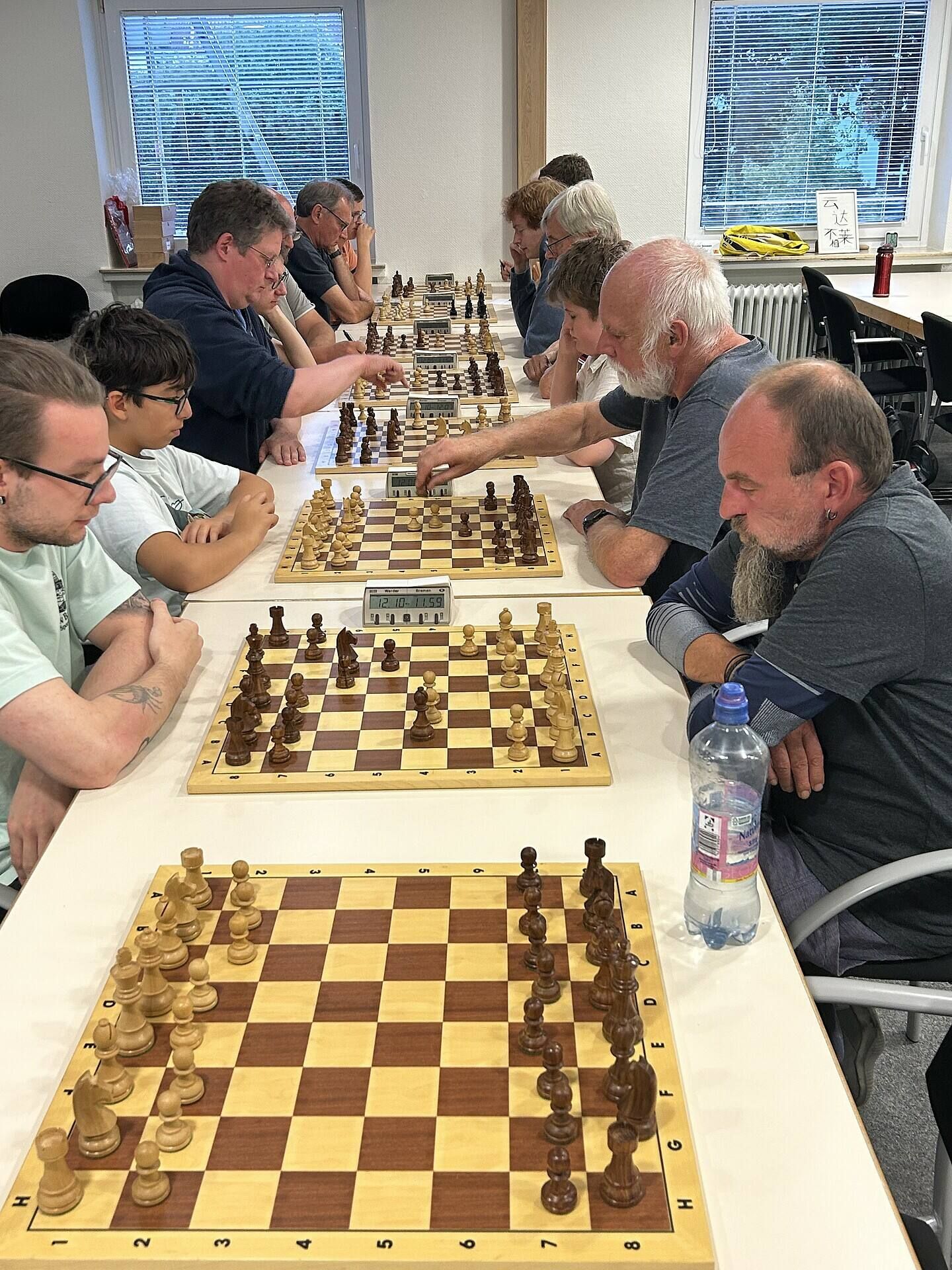 Menschen unterschiedlichen Alters spielen zu zweit Schach an einem langen Tisch in einem gut beleuchteten Raum und konzentrieren sich auf ihre Partien.