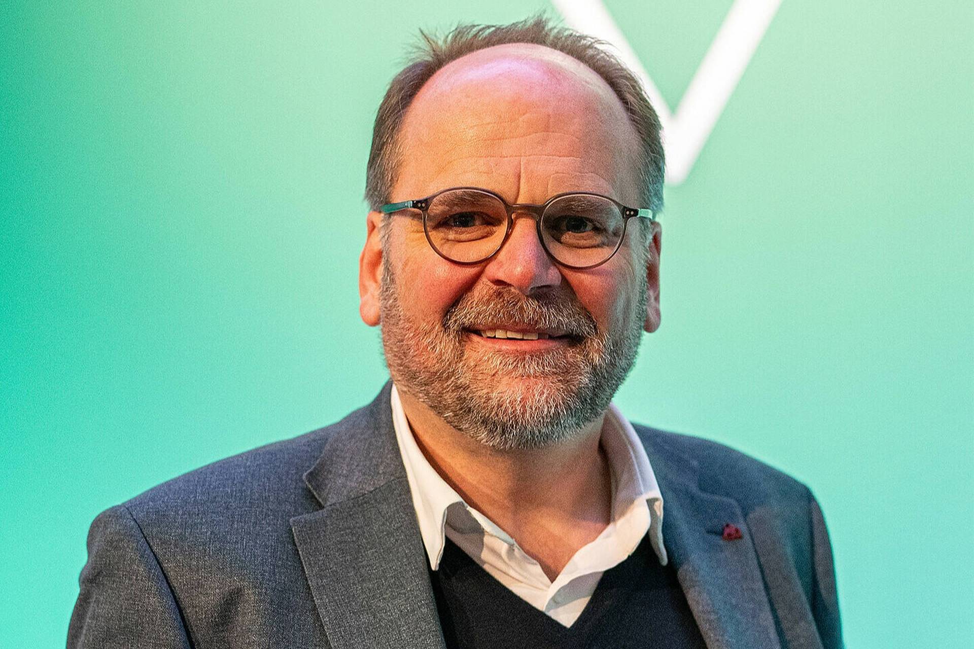 Lächelnder Mann mit Brille und Bart, bekleidet mit Anzug und Pullover, steht vor einem grünen Hintergrund.
