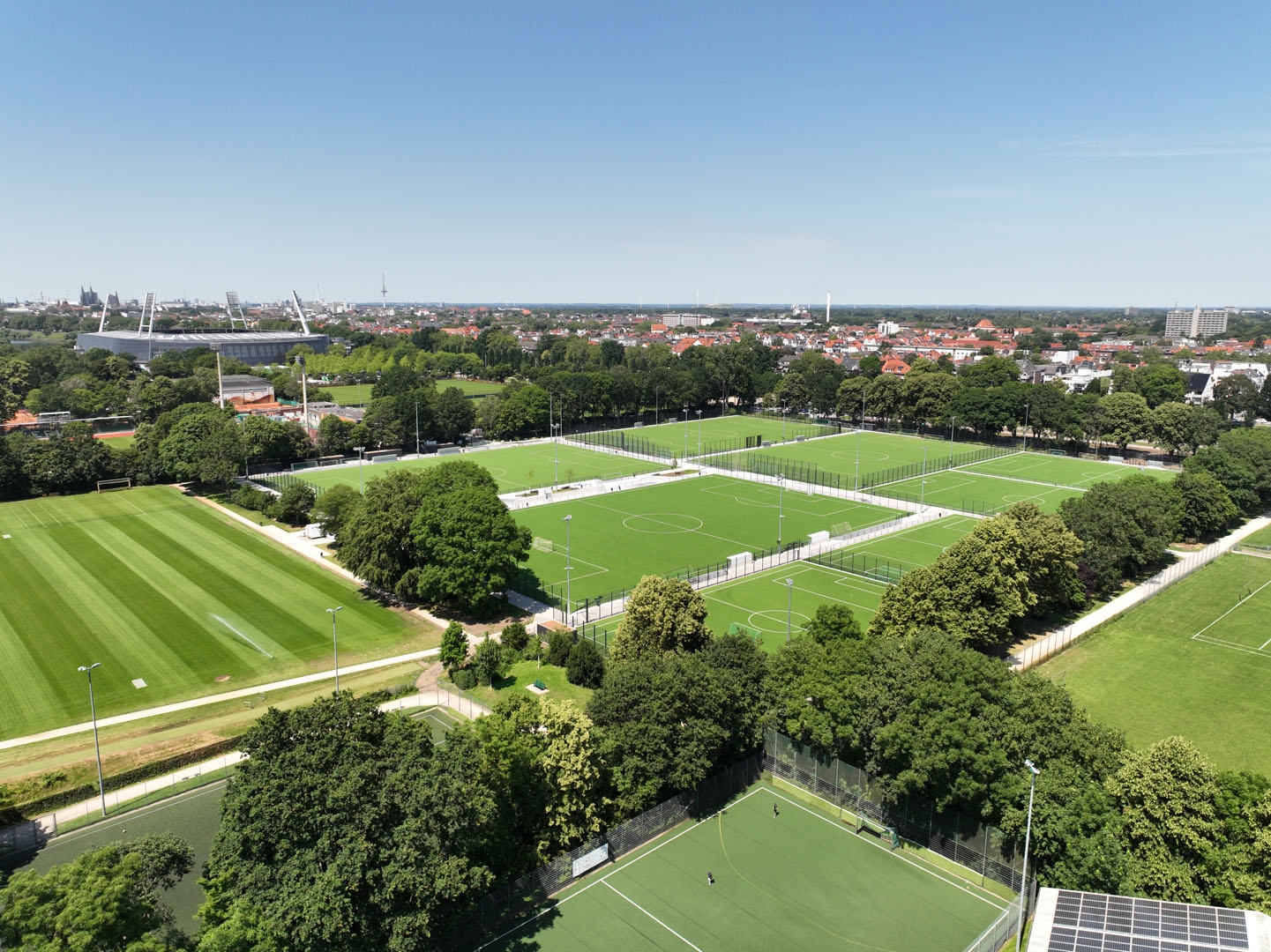 Luftaufnahme zahlreicher nebeneinander liegender, von Bäumen eingerahmter Fußballplätze, im Hintergrund sind unter klarem Himmel die Skyline und ein Stadion zu sehen.