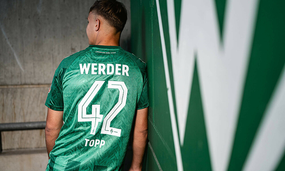Keke Topp trägt das neue, grüne Werder-Trikot. Er steht angelehnt an eine Wand. Man sieht seinen Rücken. Darauf ist die Nummer 42.