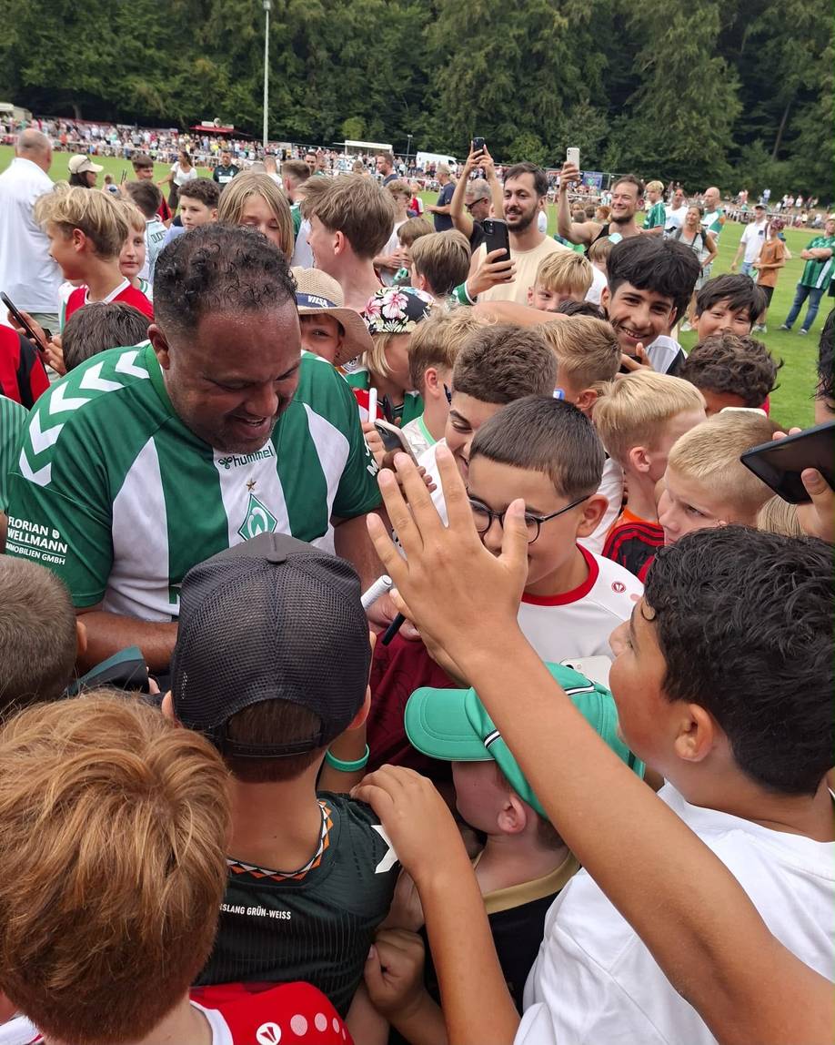 Ailton schreibt Autogramme auf dem Platz. Er ist umringt von Fans.