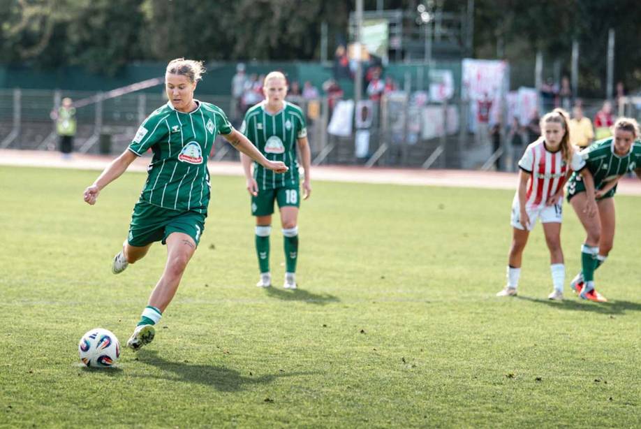 Fußballerin in grünem Trikot schießt den Ball auf einem Rasenplatz, Teamkolleginnen und Gegnerinnen schauen zu.