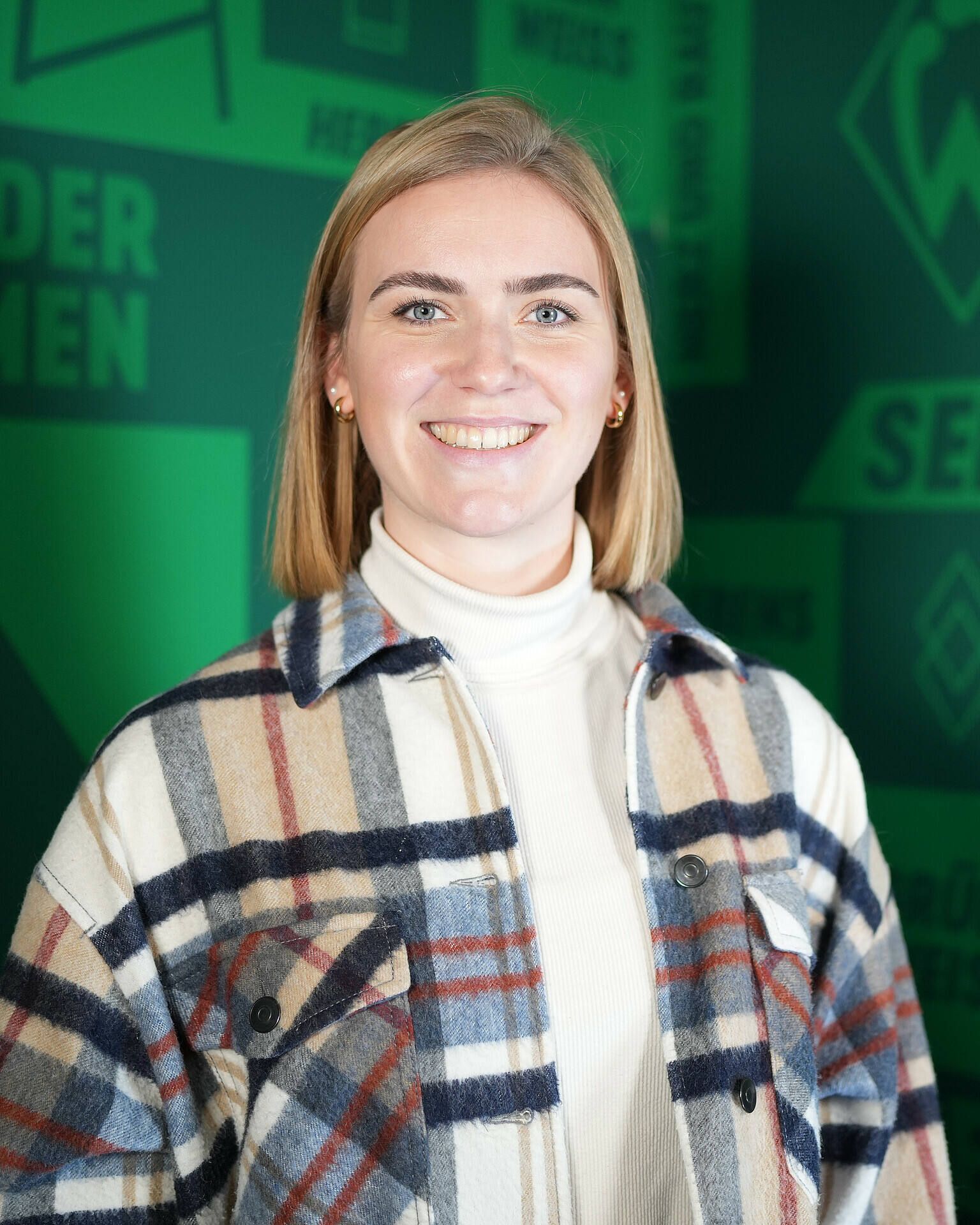 Lächelnde Person mit schulterlangen Haaren, die eine karierte Jacke und einen weißen Rollkragenpullover trägt, vor einem grünen Hintergrund mit Text.