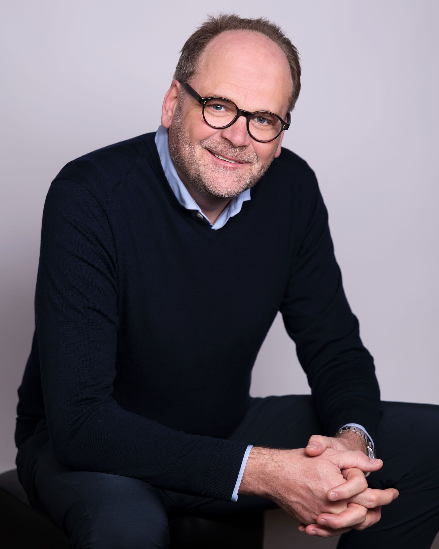 Ein lächelnder Mann mit Brille und Bart, der einen dunkelblauen Pullover trägt, sitzt mit gefalteten Händen vor einem schlichten Hintergrund.