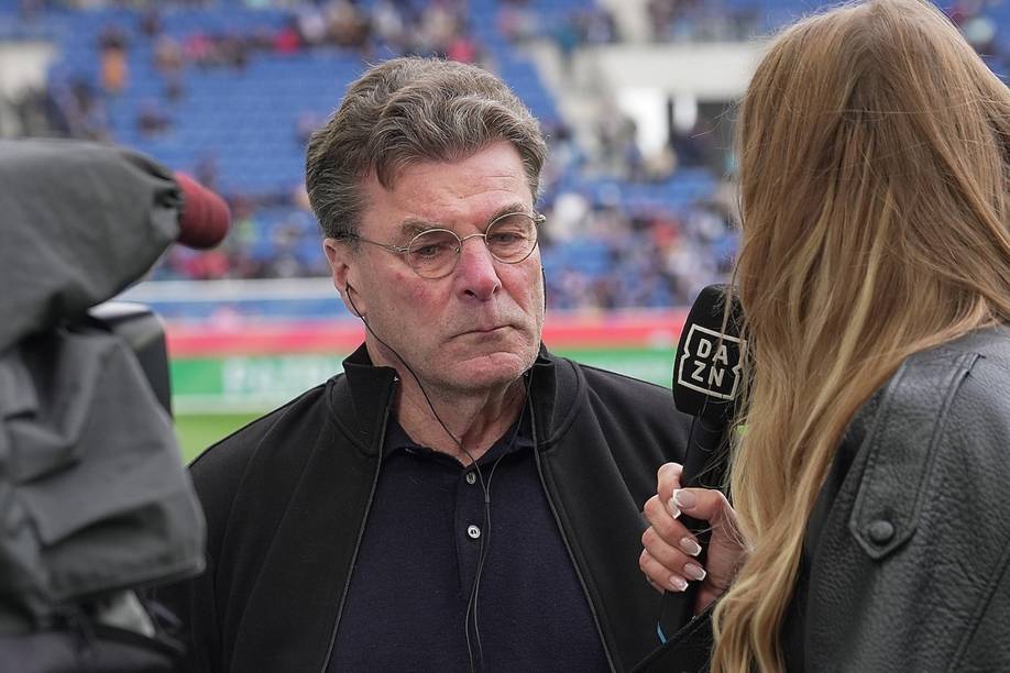 Ein älterer Mann mit Brille wird in einem Sportstadion interviewt. Der Interviewer hält ein Mikrofon mit der Aufschrift „DAZN“ in der Hand.