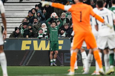 Olivier Deman steht an der Seitenlinie vor einer Werbebande. Er trägt ein grünes Trikot, grüne Hose und grüne Stutzen. Er hält einen weiß-bunten Ball mit beiden Händen über seinem Kopf. Im Vordergrund sind unscharf andere Spieler in weiß und orange zu sehen.