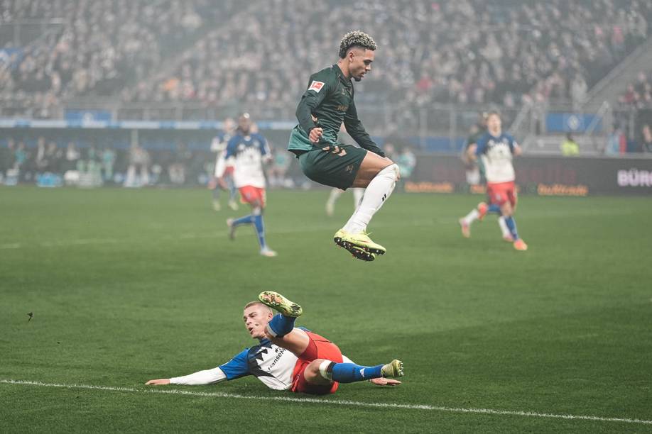 Ein Fußballspieler springt während eines Spiels über einen sich hinwerfenden Gegner hinweg, im Hintergrund ist eine verschwommene Menschenmenge zu sehen.