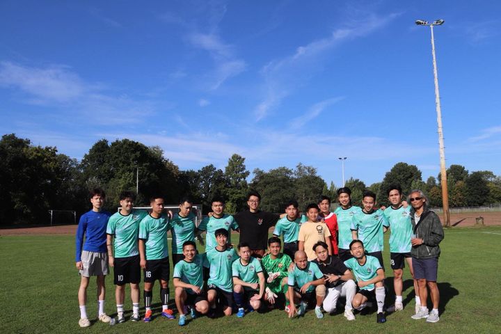 Eine Fußballmannschaft posiert auf einem Feld unter strahlend blauem Himmel in grünen Trikots, im Hintergrund sind Bäume und ein Lichtmast zu sehen.