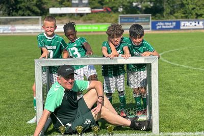 Auf einem sonnigen Spielfeld sitzt ein Trainer lächelnd vor einem kleinen Fußballtor, hinter dem vier junge Spieler in grünen Trikots stehen.