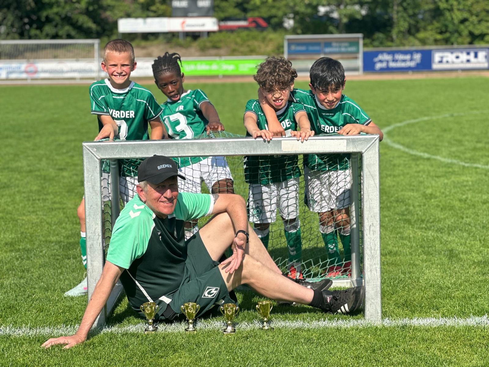 Auf einem sonnigen Spielfeld sitzt ein Trainer lächelnd vor einem kleinen Fußballtor, hinter dem vier junge Spieler in grünen Trikots stehen.