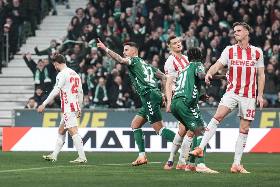 Fußballspielszene: Ein Spieler in Grün hat den Zeigefinger ausgestreckt, weitere Spieler in weißen Trikots mit roten Streifen reagieren emotional. Im Hintergrund jubelt die Menge im Stadion.