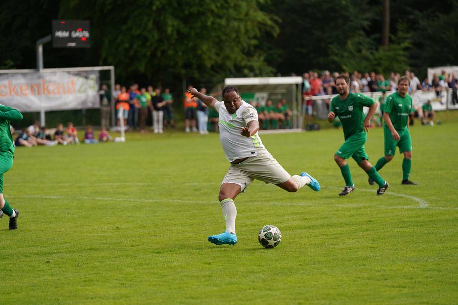 Ein Fußballspieler in weißer Spielkleidung macht sich auf einem Rasenfeld bereit, den Ball zu schießen, während Gegner in grünen Trikots von hinten herankommen.