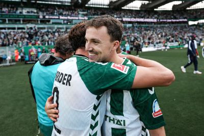 Zwei Fußballspieler in grün-weißen Trikots umarmen sich lächelnd auf dem Spielfeld, im Hintergrund jubelt die Menge.