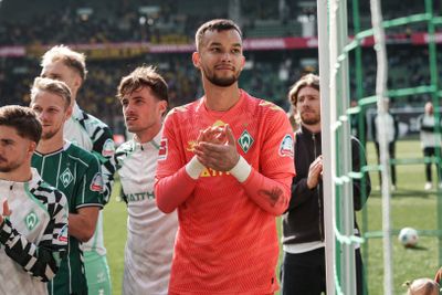 Fußballspieler in Mannschaftstrikots klatschen und stehen auf einem Spielfeld, im Hintergrund ist eine Menschenmenge zu sehen.