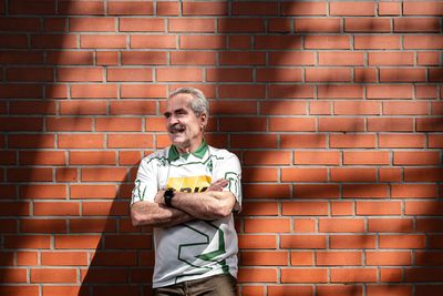 Ein Mann in einem weißen Sporttrikot lehnt mit verschränkten Armen an einer Backsteinmauer, neben ihm auf dem Boden steht eine silberne Trophäe.