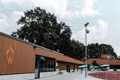 Moderne Sportanlage mit Holzfassade, angrenzender Laufbahn und vereinzelten Bäumen unter einem teilweise bewölkten Himmel.