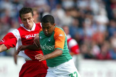 Zwei Fußballspieler kämpfen während eines Spiels um den Ball, einer im rot-weißen Trikot, der andere im grün-orangefarbenen, im Hintergrund ist eine verschwommene Menschenmenge zu sehen.