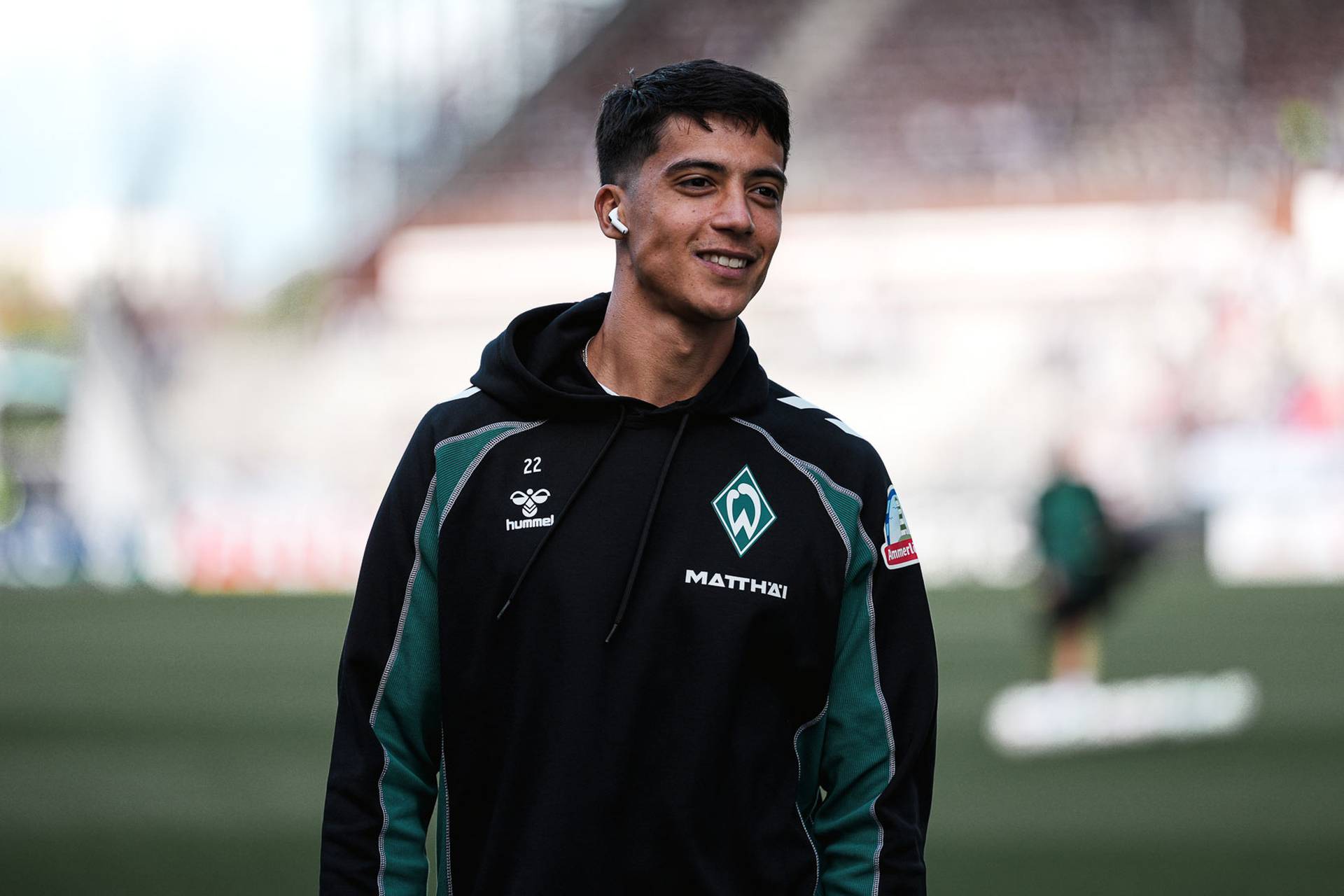 Auf einem Fußballfeld lächelt eine Person, die eine schwarz-grüne Jacke mit der Nummer 22 und einem Vereinslogo trägt und Kopfhörer in den Ohren hat.