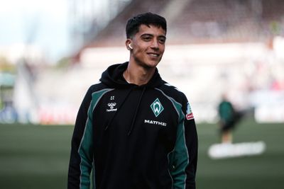 Auf einem Fußballfeld lächelt eine Person, die eine schwarz-grüne Jacke mit der Nummer 22 und einem Vereinslogo trägt und Kopfhörer in den Ohren hat.