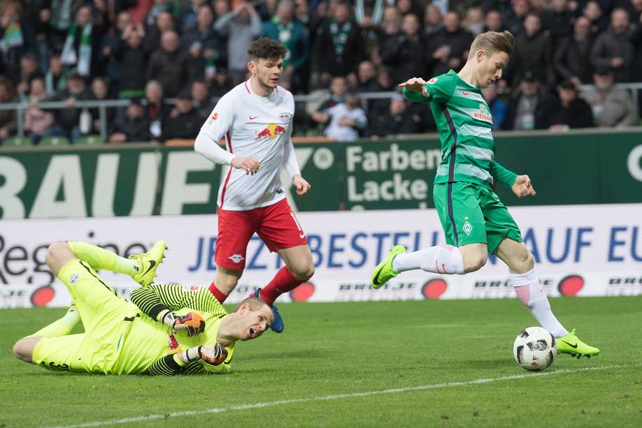 Ein Fußballspieler im grünen Trikot versucht einen Schuss, während ein Torwart in neonfarbener Uniform sich hinwirft, um den Ball abzuwehren; im Hintergrund sind Zuschauer zu sehen.