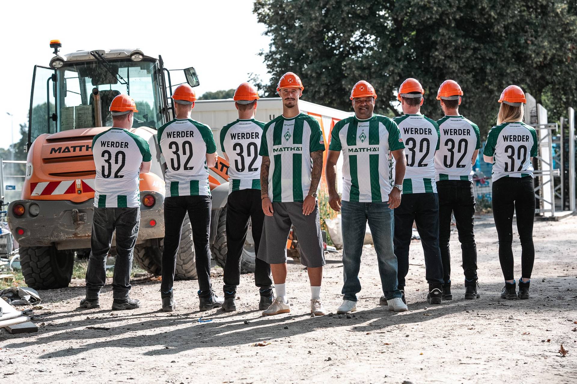 Sieben Personen in Werder-Trikots und mit Schutzhelmen stehen an einem sonnigen Tag in der Nähe von Baumaschinen.