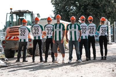 Sieben Personen in Werder-Trikots und mit Schutzhelmen stehen an einem sonnigen Tag in der Nähe von Baumaschinen.