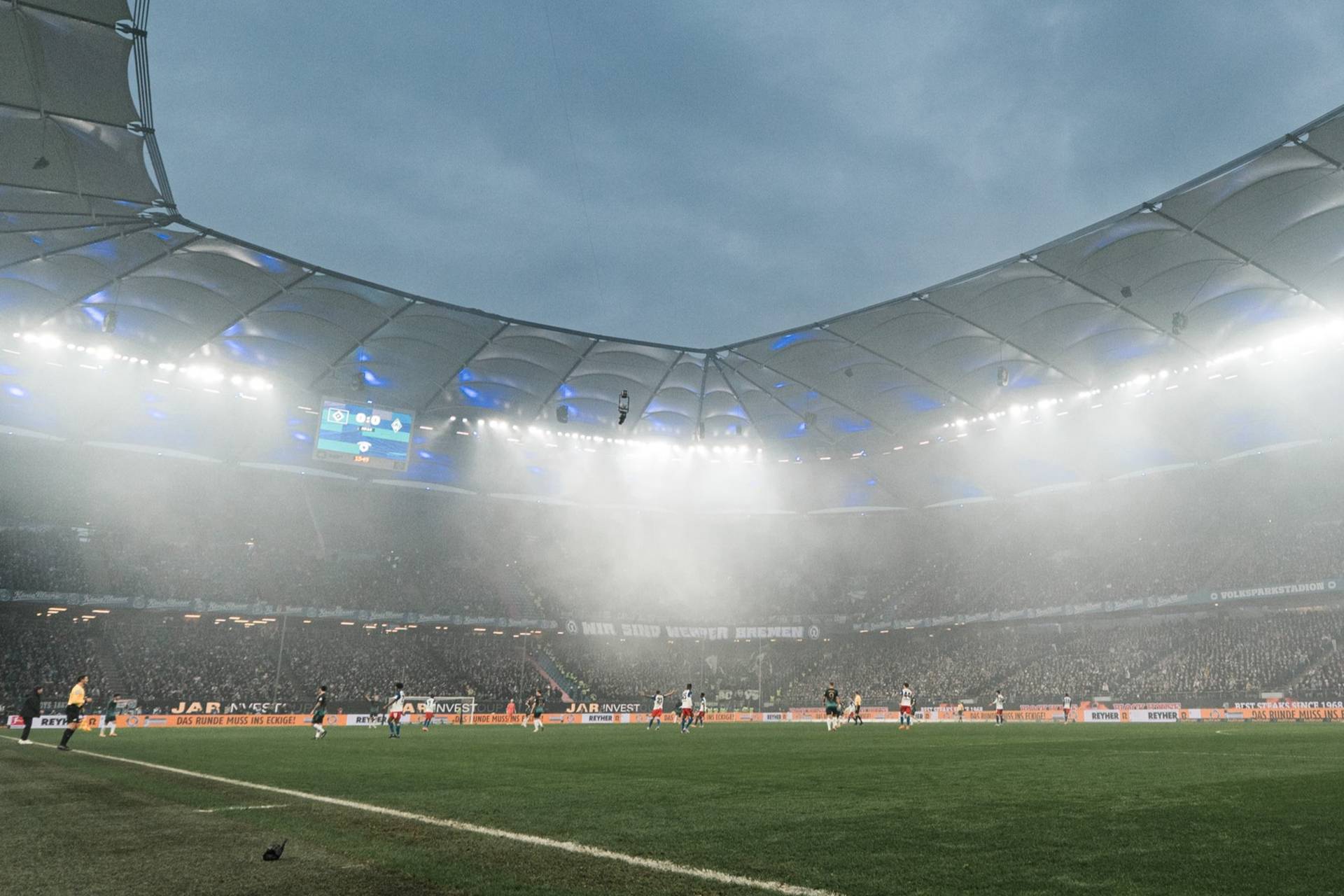 Fußballspiel in einem großen, nebligen Stadion mit hellen Lichtern und teilweise gefüllten Zuschauerrängen unter bewölktem Himmel.