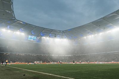 Fußballspiel in einem großen, nebligen Stadion mit hellen Lichtern und teilweise gefüllten Zuschauerrängen unter bewölktem Himmel.