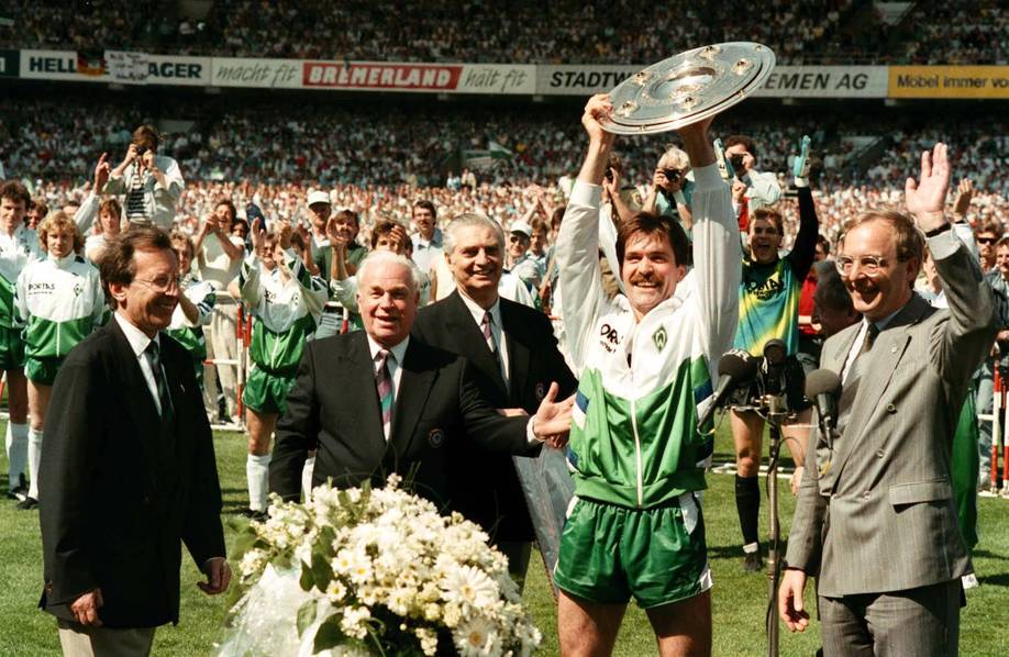 Ein Fußballspieler in einem grünen Trainingsanzug hält auf dem Spielfeld einen Pokal in die Höhe, umgeben von applaudierenden Offiziellen und Mannschaftskameraden, im Hintergrund ist eine Menschenmenge zu sehen.