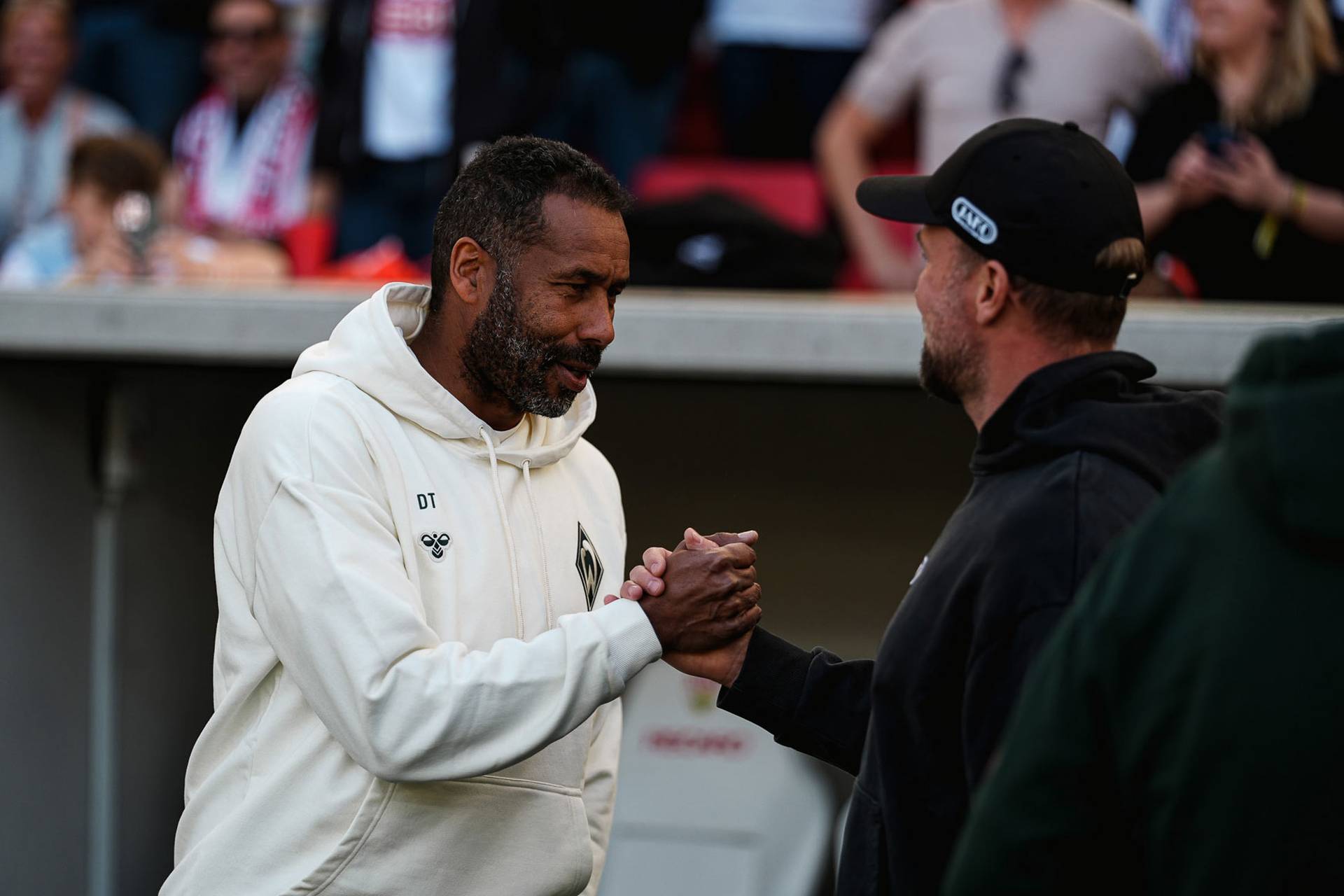 Zwei Männer, die sich bei einer Veranstaltung im Freien die Hand geben; der eine trägt einen weißen Kapuzenpulli, der andere eine schwarze Mütze. Im unscharfen Hintergrund ist eine Menschenmenge zu sehen.