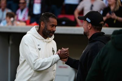 Zwei Männer, die sich bei einer Veranstaltung im Freien die Hand geben; der eine trägt einen weißen Kapuzenpulli, der andere eine schwarze Mütze. Im unscharfen Hintergrund ist eine Menschenmenge zu sehen.