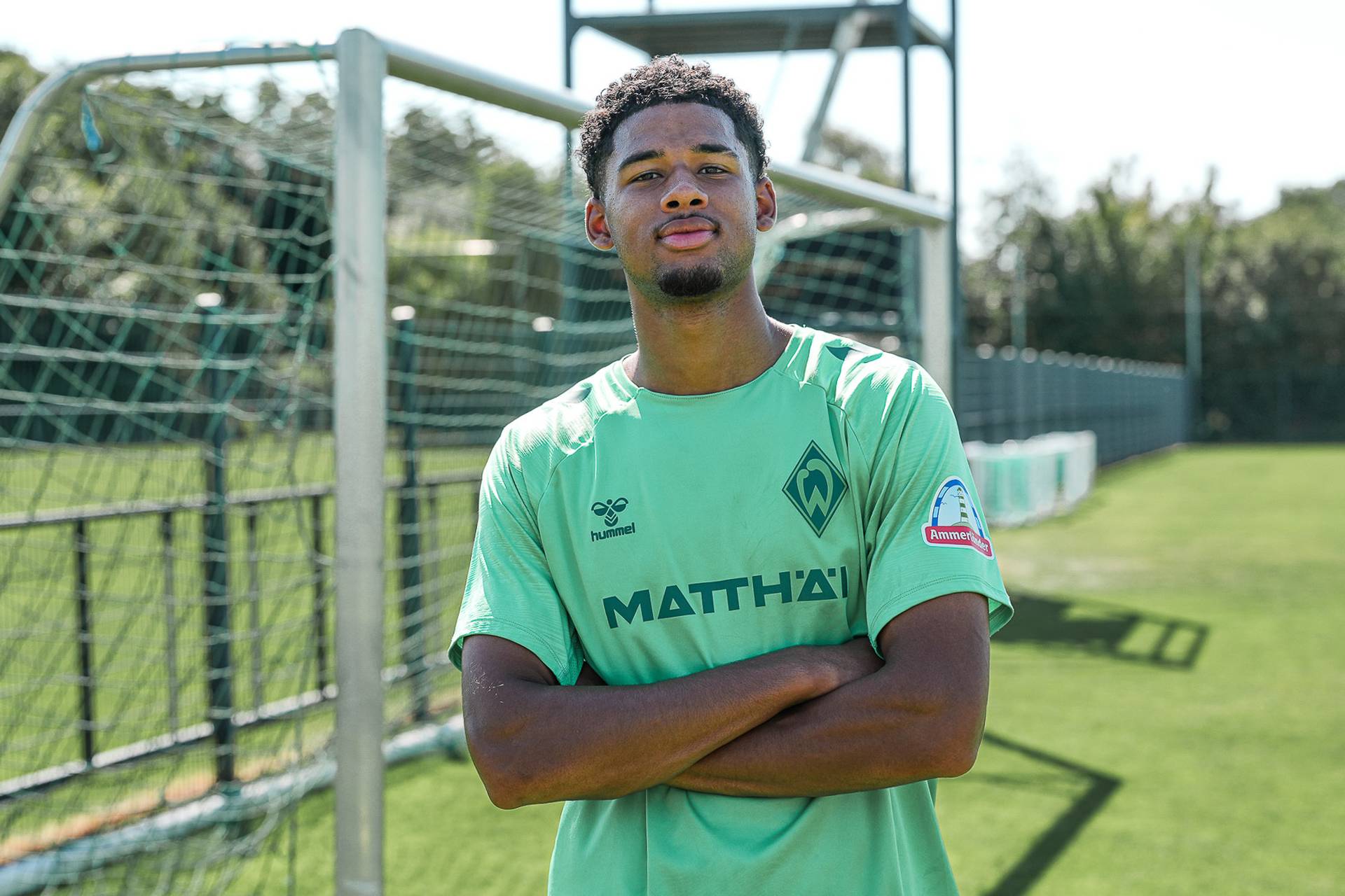 Ein Fußballspieler im grünen Trikot von Werder Bremen steht mit verschränkten Armen auf einem Fußballplatz, im Hintergrund ist ein Tor zu sehen.