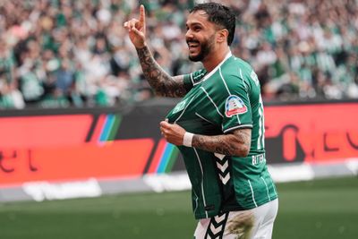 Ein Fußballspieler im grünen Trikot feiert ein Tor, zeigt lächelnd nach oben, im Hintergrund ist ein Stadion mit einer verschwommenen Menschenmenge zu sehen.