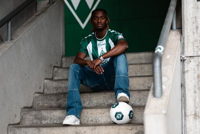Eine Person sitzt auf Betonstufen, bekleidet mit einem grün-weißen Trikot, Jeans und weißen Turnschuhen, neben ihr liegt ein Fußball, im Hintergrund ist eine grüne Wand zu sehen.