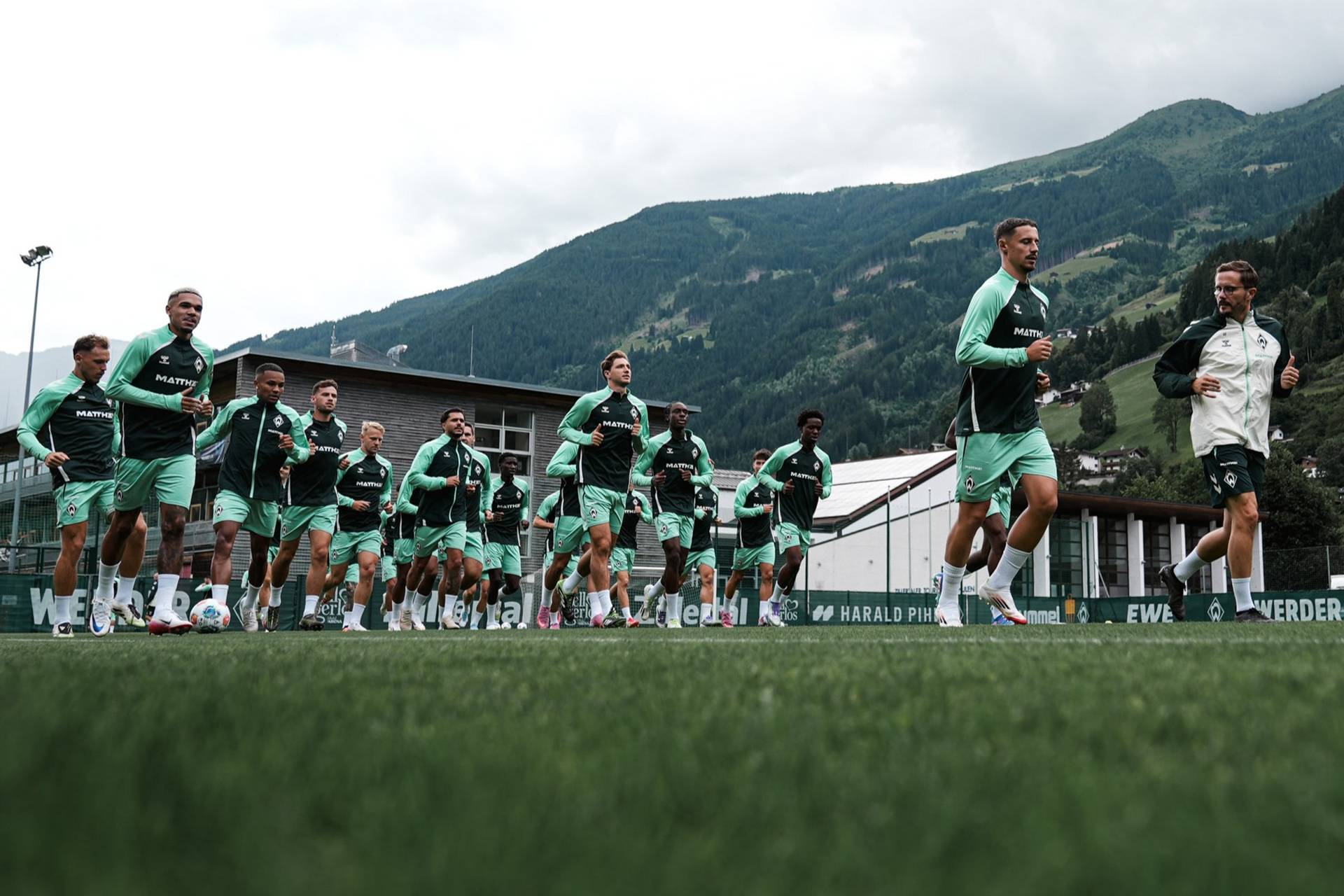 Fußballmannschaft in grünen Trainingsanzügen joggt über ein Feld mit alpinen Bergen und Gebäuden im Hintergrund.
