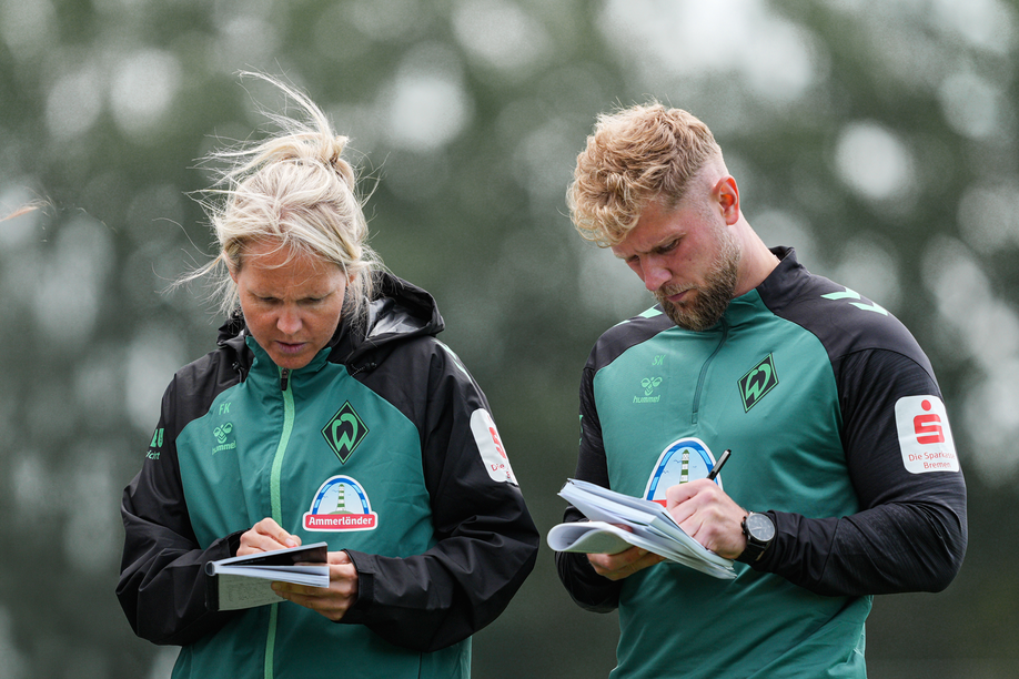 Zwei Personen in Werder-Bremen-Sportjacken konzentrieren sich darauf, im Freien Notizen zu machen; sie halten Notizbücher und Stifte in der Hand, vor einem verschwommenen, von Blättern geprägten Hintergrund.