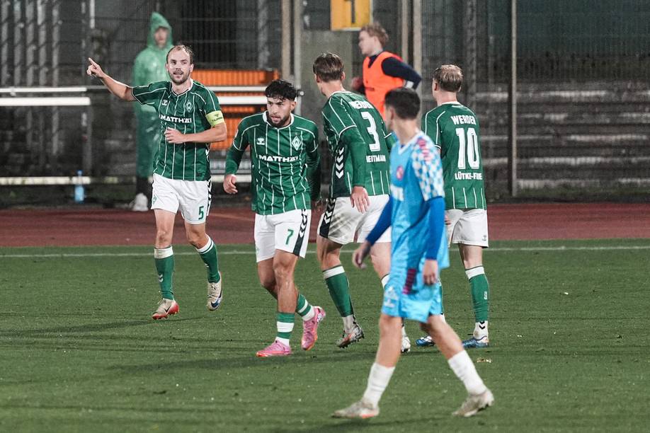 Fußballspieler in grünen Trikots feiern auf einem regennassen Spielfeld ein Tor, während ihre Gegner in blauen und Trikots stehen.